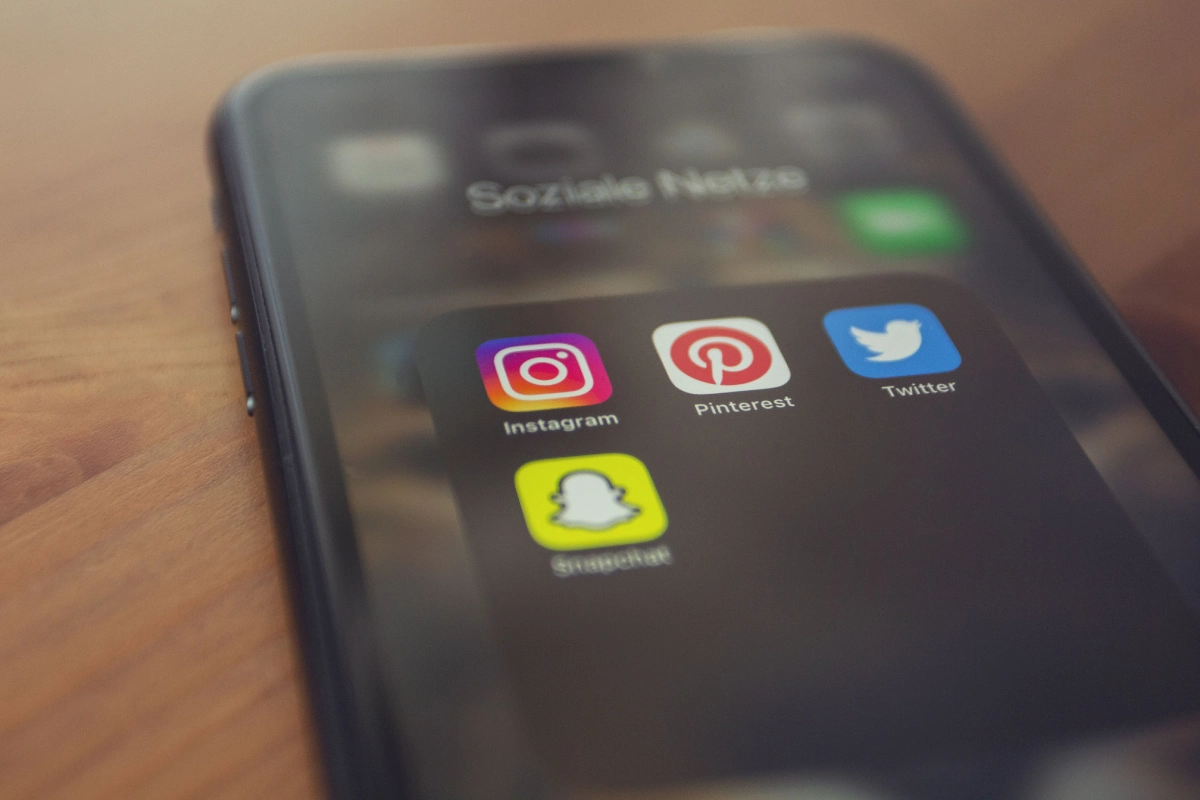 Pantalla de un teléfono inteligente recostado sobre una mesa de madera, mostrando una carpeta de aplicaciones de redes sociales como Instagram, Pinterest, Twitter y Snapchat.