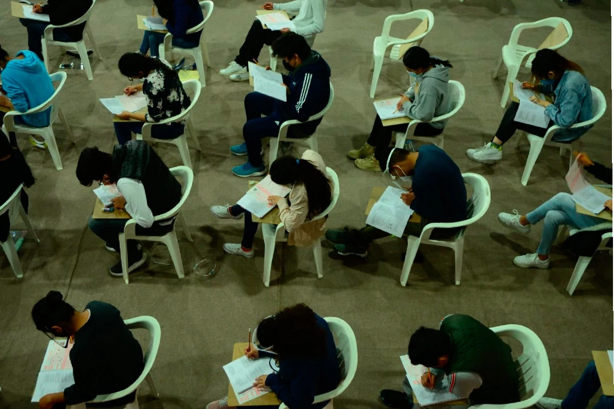 ALumnos acomodados por fila sentados en sillas blancas y con una tabla de madera para apoyarse al contestar su examen 