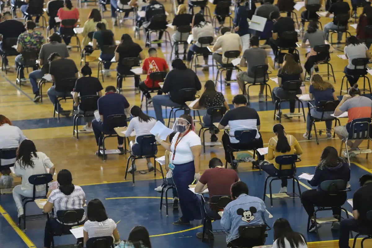Vista elevada de cientos de aspirantes realizando un examen en pupitres separados dentro de un gimnasio o auditorio, mientras una aplicadora supervisa la prueba.