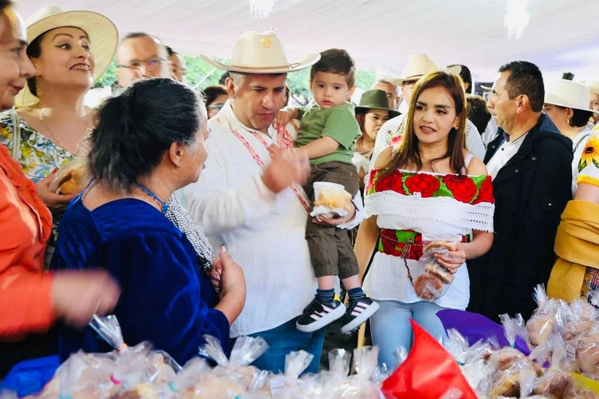 Carlos Manzo con sombrero vaquero cargando a un niño pequeño mientras conversan con locatarios y acompañan a Grecia Quiroz, quien luce una blusa con bordados florales tradicionales, en un evento público rodeado de productos artesanales.