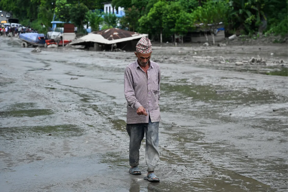 Hombre mayor nepalí con gorro tradicional dhaka topi caminando cabizbajo sobre una calle cubierta de lodo y agua tras una avalancha o inundación.