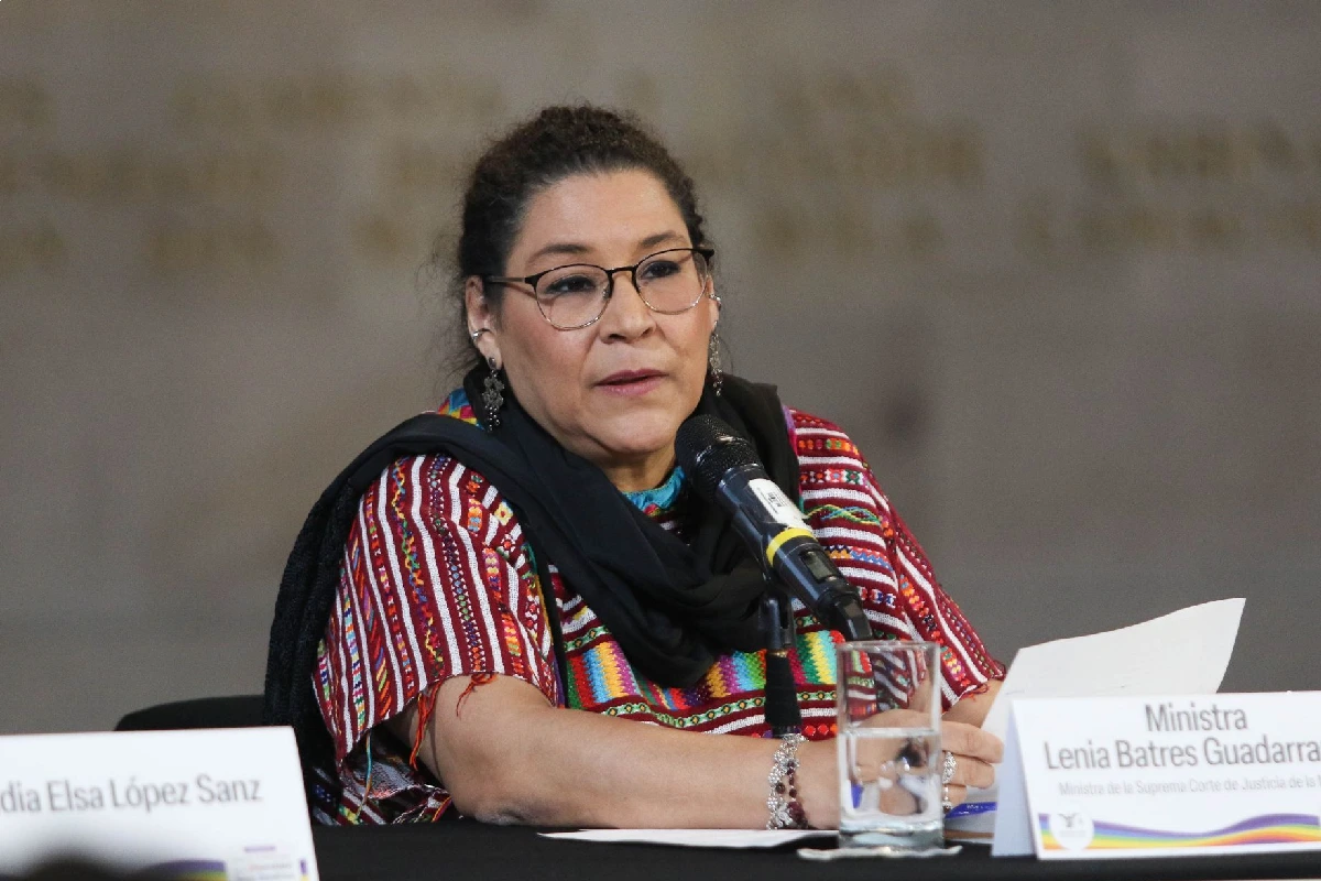 La ministra Lenia Batres Guadarrama vistiendo un huipil multicolor y anteojos, hablando frente a un micrófono en una mesa de presídium con su identificador oficial.
