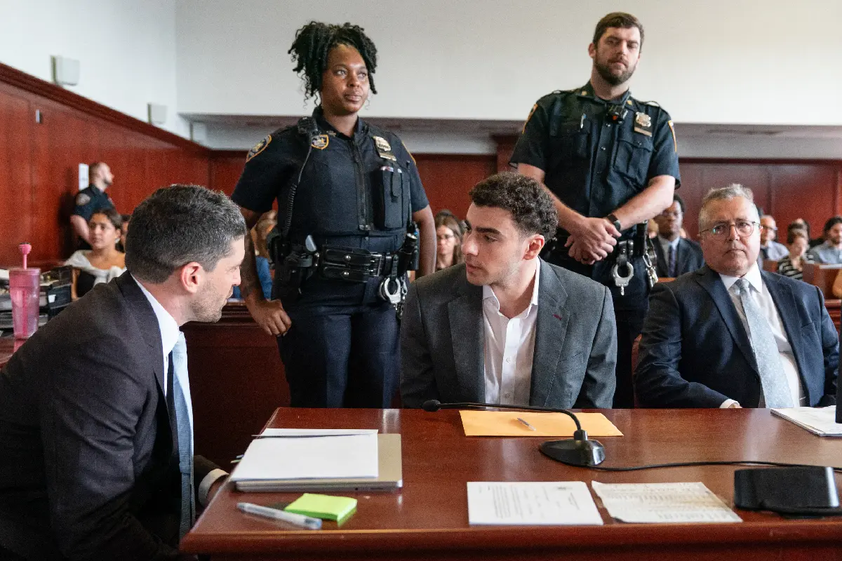 Luigi Mangione sentado en la mesa de la defensa en un tribunal, hablando con su abogado mientras dos oficiales de policía de Nueva York permanecen de pie detrás de ellos.