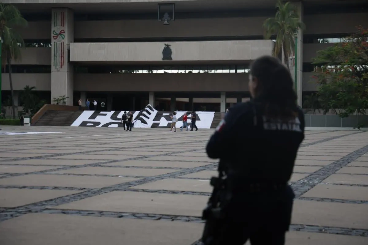 Elemento de la Policía Estatal de espaldas observando desde la explanada el Palacio de Gobierno de Sinaloa, donde manifestantes sostienen una manta con la leyenda "FUERA ROCHA" en las escalinatas.