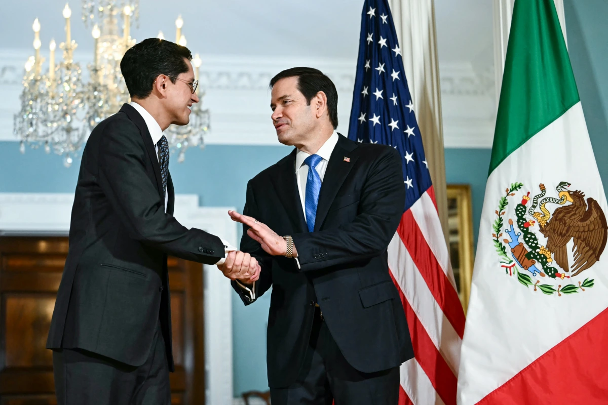 Roberto Velasco y Marco Rubio estrechándose las manos en un apretón diplomático, vestidos de traje y frente a las banderas oficiales de Estados Unidos y México.