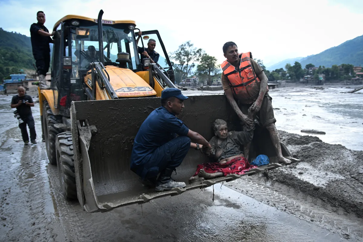 Rescatistas en Nepal auxilian a una mujer mayor cubierta de lodo transportándola en la pala de una retroexcavadora en una zona devastada por inundaciones.