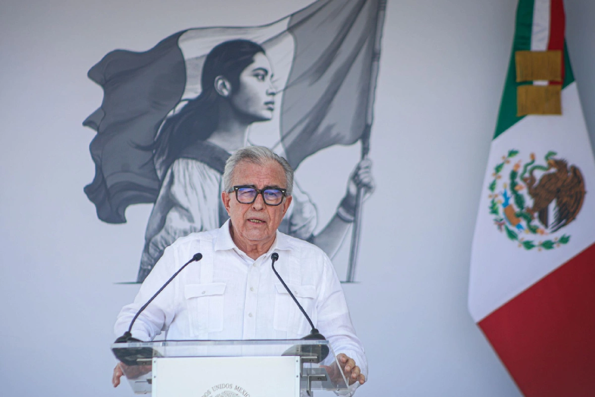 Rubén Rocha Moya con guayabera blanca y anteojos habla en el pódium institucional, con la bandera de México a su lado y el logotipo del Gobierno Federal al fondo.