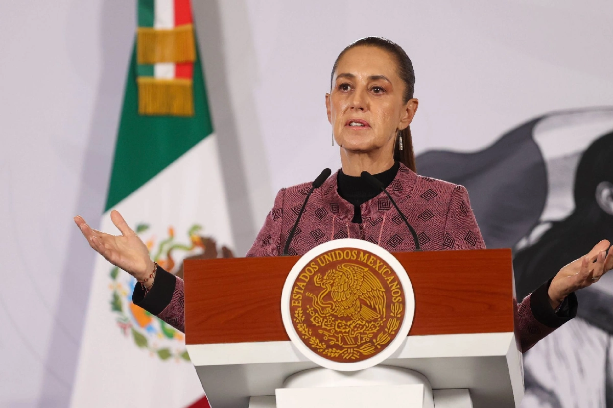 La presidenta Claudia Sheinbaum hablando en el pódium oficial con el Escudo Nacional, vistiendo un saco rosa con patrones geométricos y manteniendo los brazos abiertos con las palmas arriba.