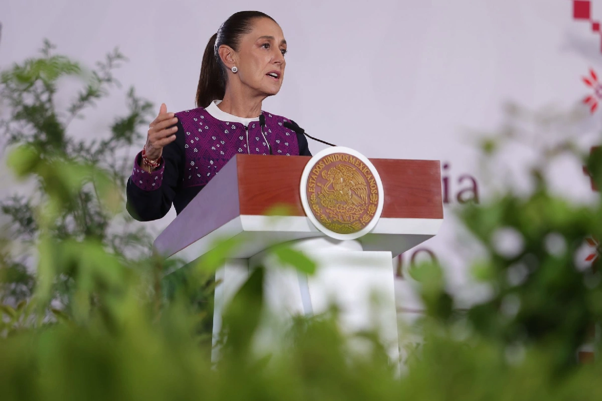 Claudia Sheinbaum de perfil detrás de un pódium con el escudo de México, vistiendo una prenda con bordados morados y gesticulando mientras habla, enmarcada por plantas en primer plano.