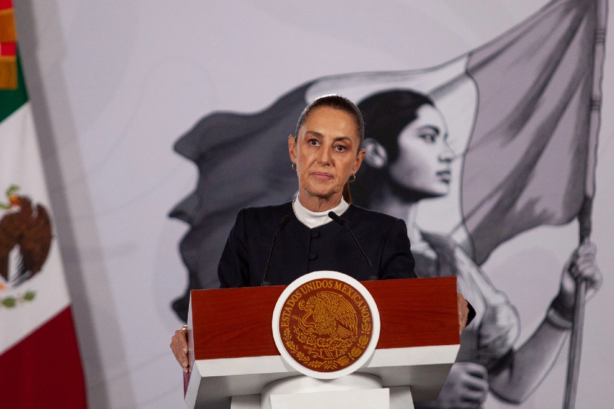 La presidenta Claudia Sheinbaum con saco oscuro en un pódium oficial con el escudo de Estados Unidos Mexicanos, flanqueada por la bandera nacional y una ilustración institucional de fondo.