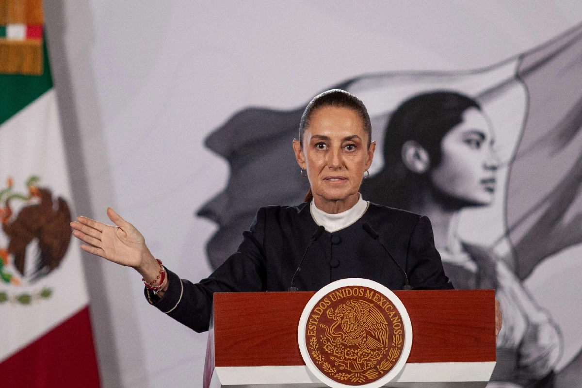 La presidenta Claudia Sheinbaum gesticulando con la mano abierta desde un pódium oficial con el escudo nacional, teniendo al fondo la bandera mexicana y un mural institucional.
