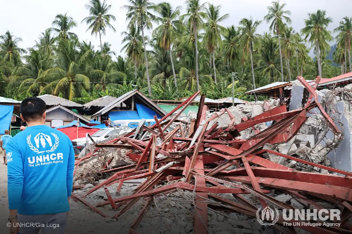 Un trabajador humanitario de ACNUR (UNHCR) con camiseta azul observa un cúmulo de vigas metálicas rojas y escombros de concreto tras un terremoto, con vegetación de palmeras al fondo.