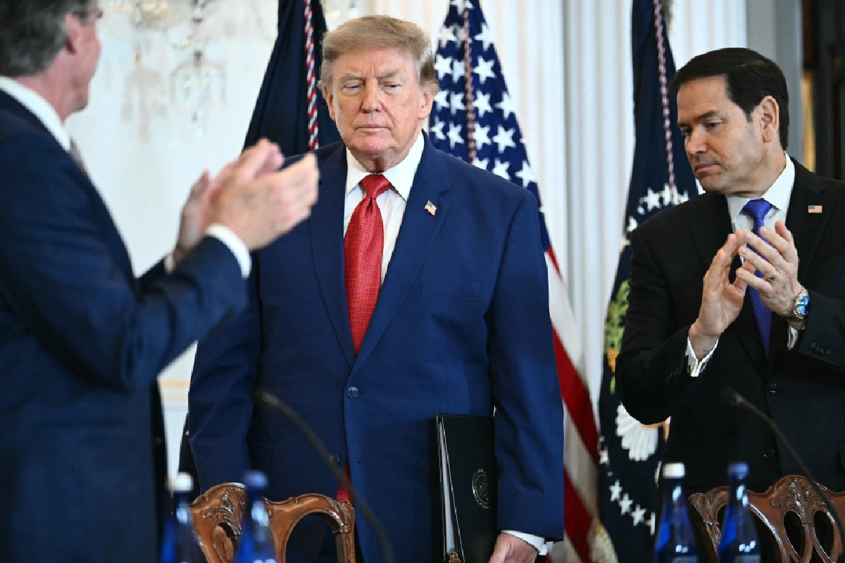 Donald Trump vistiendo un traje azul y corbata roja sostiene una carpeta mientras camina flanqueado por personas aplaudiendo, entre ellas Marco Rubio, con banderas de Estados Unidos al fondo.