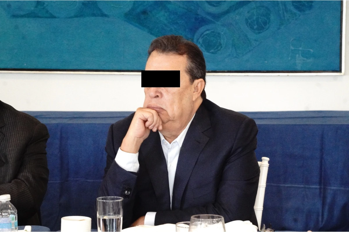 Ángel Aguirre Rivero, exgobernador de Guerrero, sentado en una mesa con el rostro parcialmente censurado por una barra negra sobre sus ojos, apoyando el mentón en su mano.