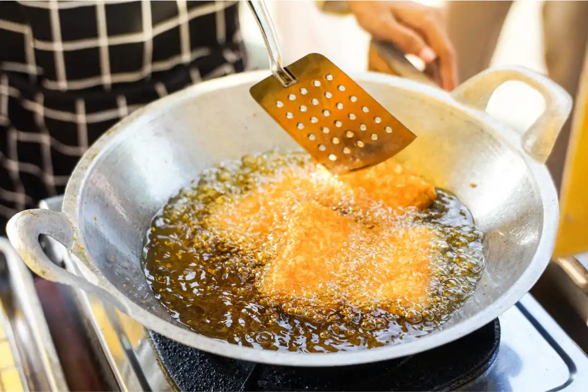 Vista de cerca de freír alimentos en aceite caliente con una espátula de metal que se utiliza, mostrando los métodos de cocina tradicionales en un entorno rústico. 