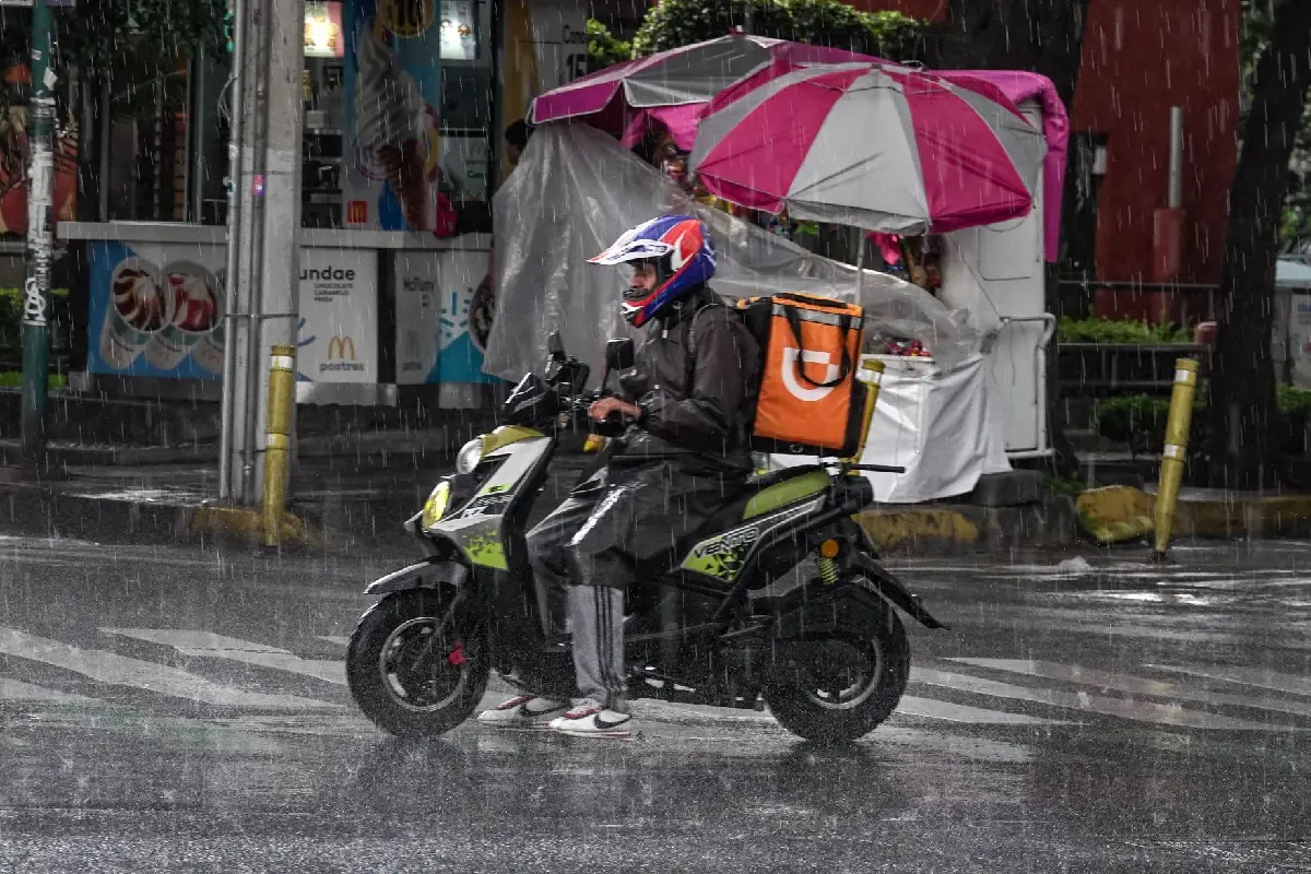 Repartidor de plataforma en motoneta con casco e impermeable conduciendo bajo una lluvia intensa en una calle de la Ciudad de México.