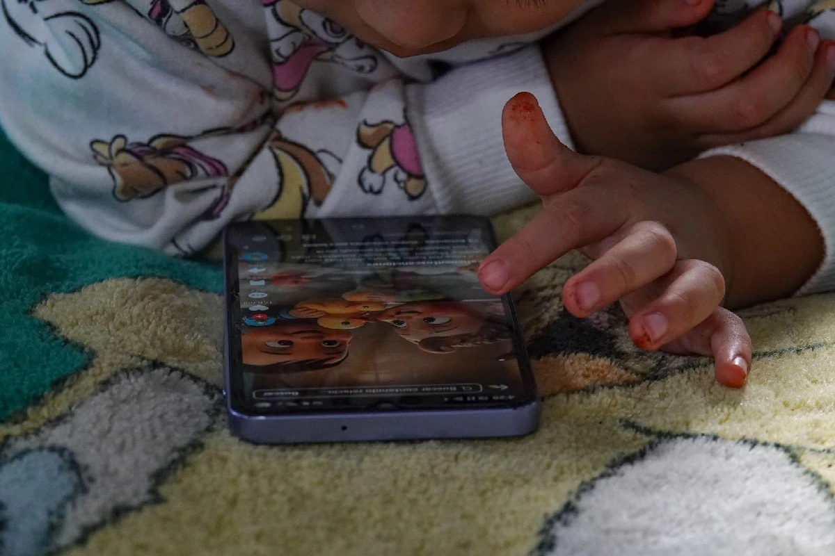 Un niño o niña recostado sobre una cobija manipulando un teléfono inteligente con el dedo en la pantalla mientras mira videos infantiles.