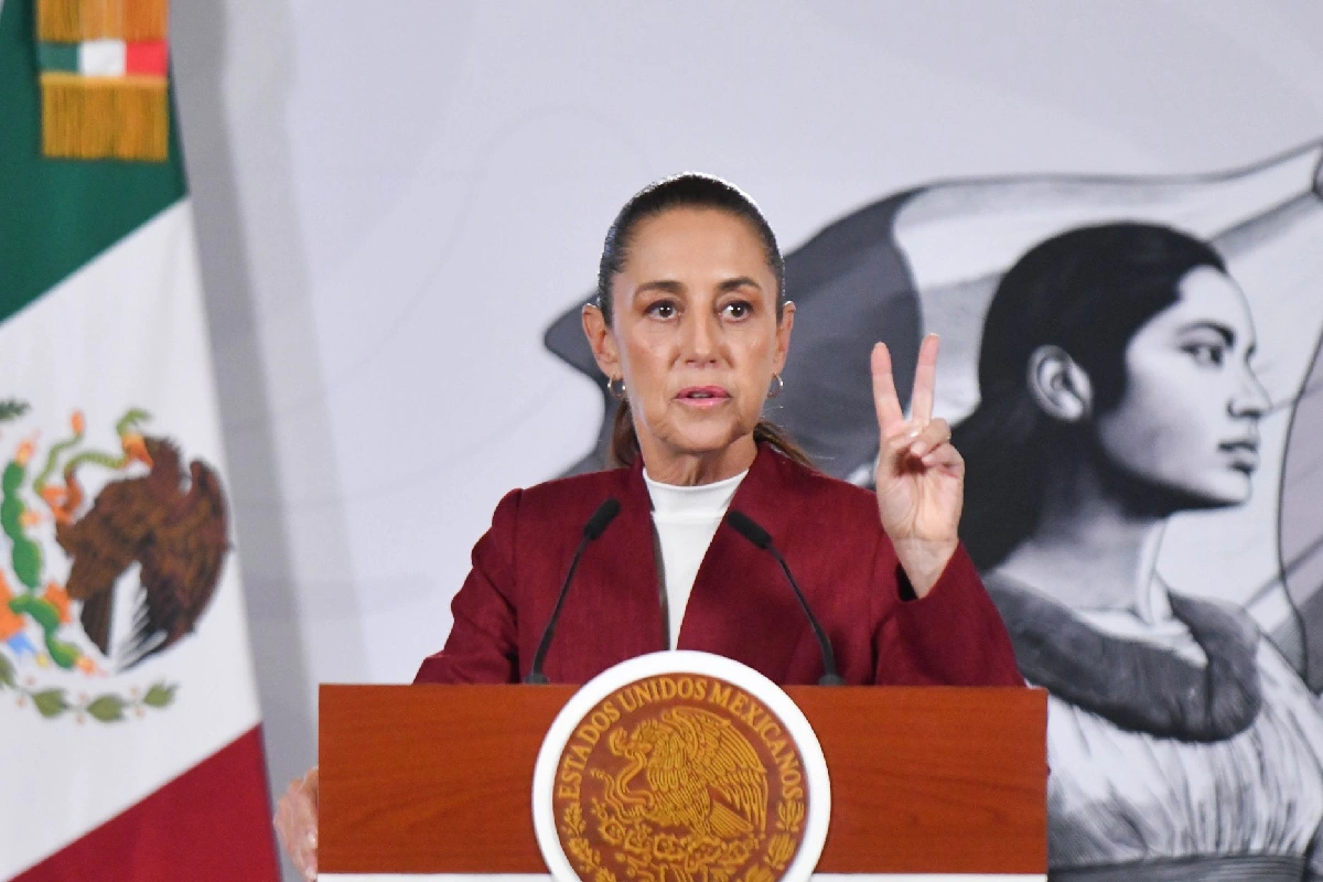 La presidenta Claudia Sheinbaum hablando desde el podio presidencial mientras levanta dos dedos durante una conferencia de prensa.