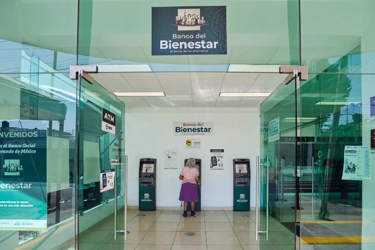 Vista frontal de la entrada a una sucursal del Banco del Bienestar con puertas de vidrio abiertas, donde una mujer adulta mayor realiza una operación en un cajero automático.