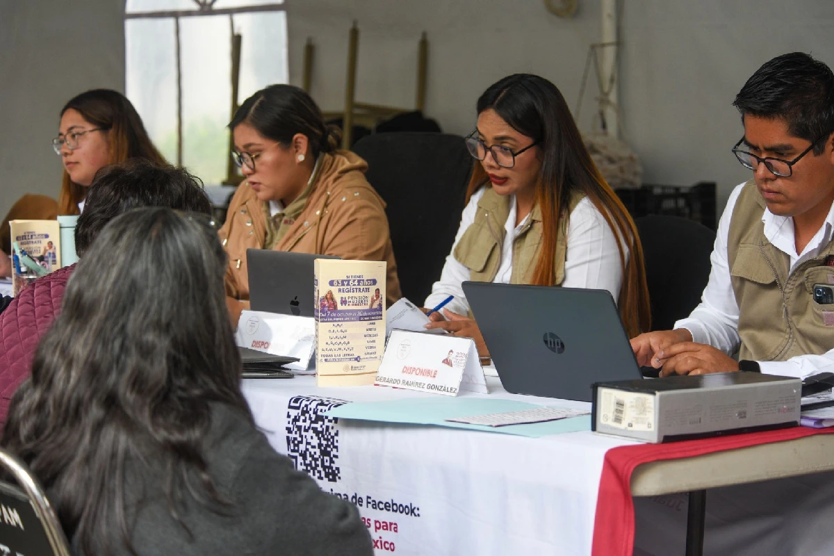 Servidores de la Nación atendiendo a ciudadanas en una mesa de registro de la Secretaría de Bienestar, rodeados de computadoras portátiles y folletos informativos sobre apoyos sociales.