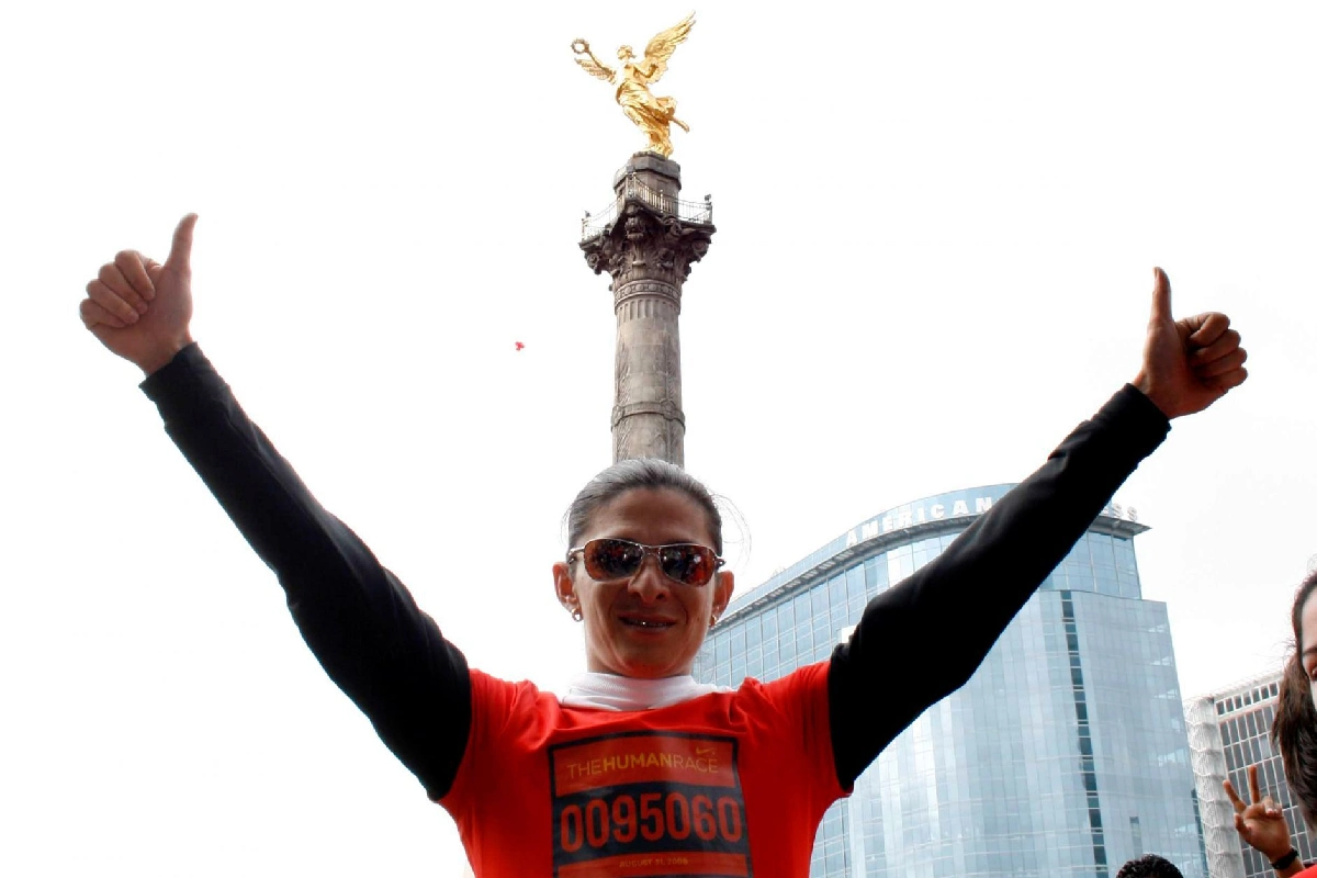 Ana Guevara levantando los brazos, usando lentes y playera deportiva con el Ángel de la Independencia detrás de ella. 