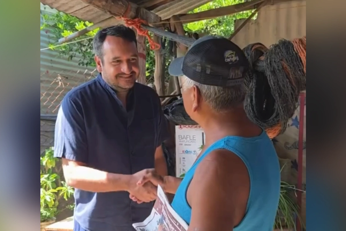 Andrés Manuel López Beltrán vistiendo camisa guayabera azul marino, sonriendo mientras estrecha la mano de un habitante local con gorra y camiseta de tirantes azul durante una visita domiciliaria.