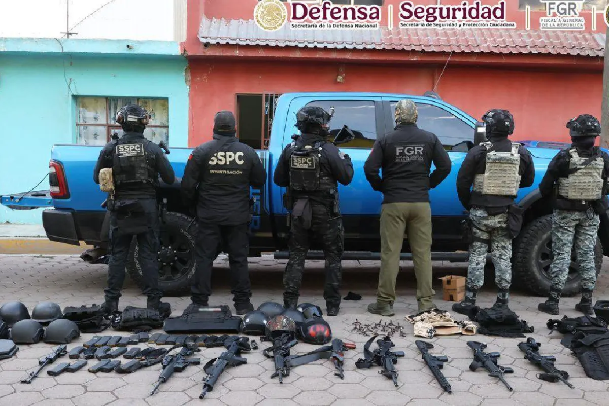 Agentes de la SSPC y FGR de espalda junto a una camioneta azul, custodiando un decomiso de armas de fuego de alto calibre, cargadores, cascos balísticos y equipo táctico desplegados sobre el suelo.