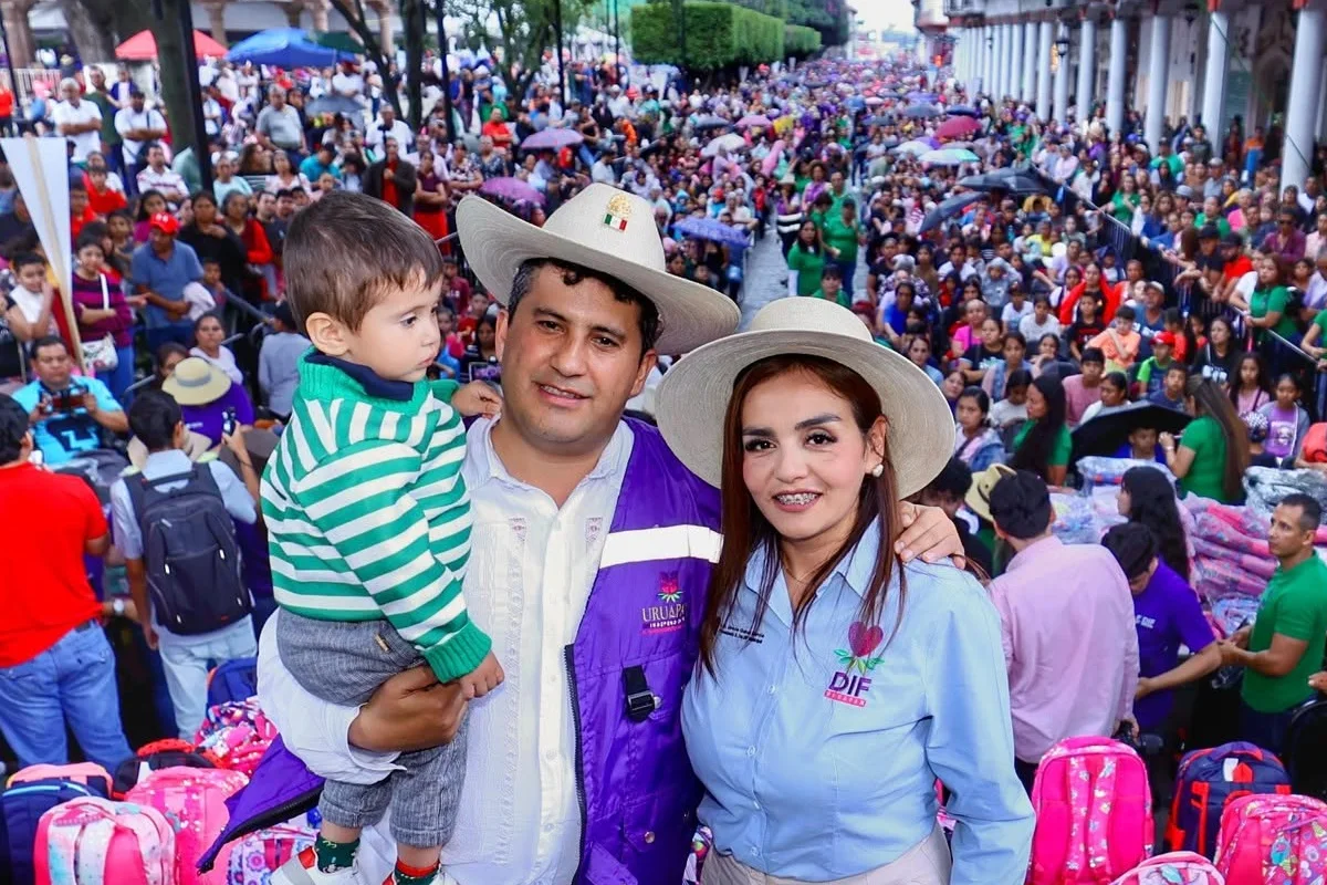 Carlos Manzo vistiendo chaleco morado y sombrero de ala ancha junto a Grecia Quiroz con camisa del DIF, sosteniendo a un niño en brazos frente a una multitud congregada en una plaza pública de Uruapan.