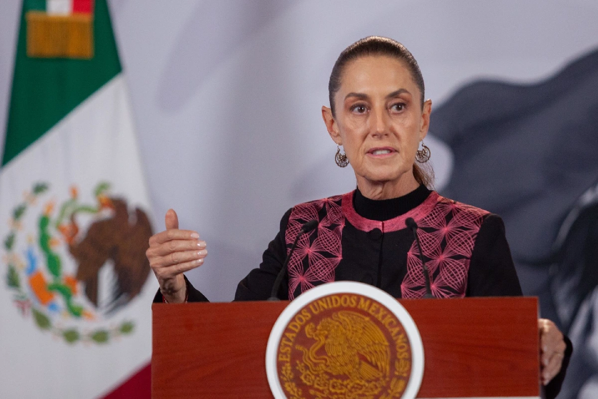Claudia Sheinbaum gesticulando con la mano derecha mientras habla en un pódium de madera con el sello oficial de Estados Unidos Mexicanos y la bandera nacional al fondo.