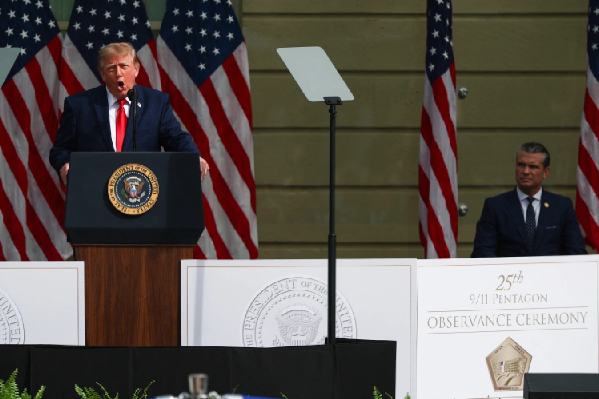 Donald Trump hablando efusivamente en un pódium presidencial con Pete Hegseth sentado a la derecha, en un escenario flanqueado por banderas estadounidenses con el letrero de la ceremonia del 25 aniversario del 11 de septiembre en el Pentágono.