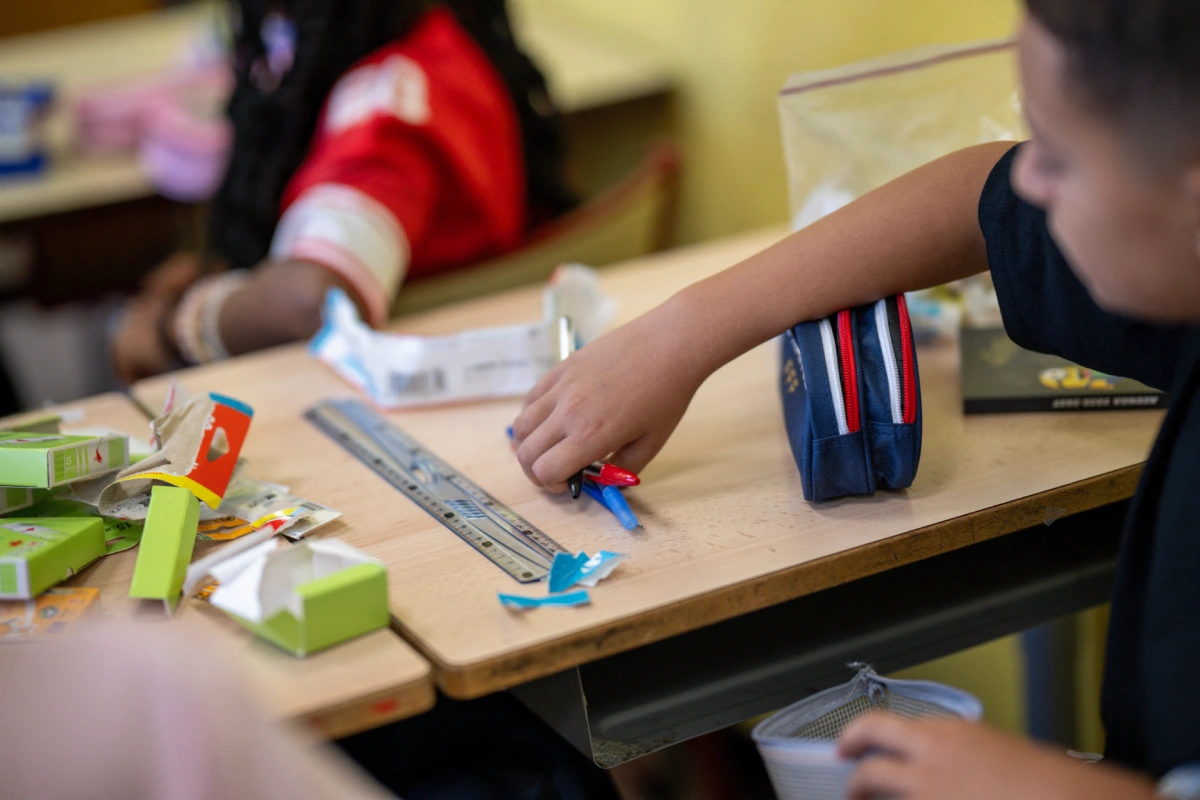 Primer plano del brazo y la mano de un estudiante tomando plumas sobre un escritorio escolar con regla, estuche y útiles educativos en un salón de clases.