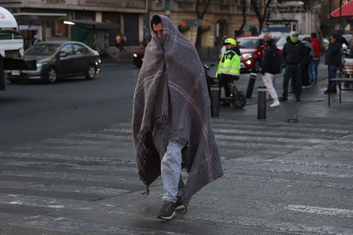 Una persona envuelta completamente en una cobija gris de lana caminando por un cruce peatonal en la calle durante un día frío en la ciudad.