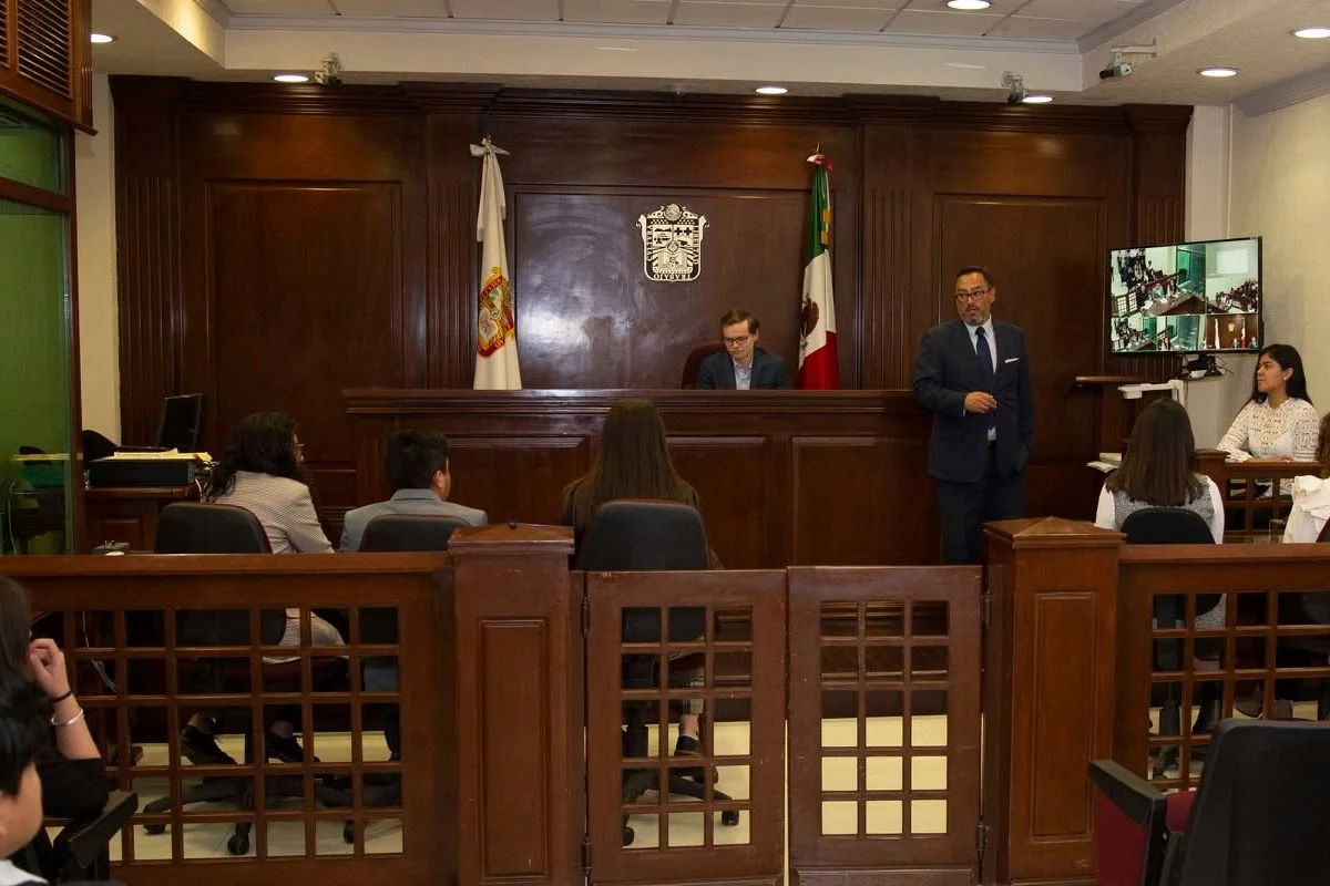 Un abogado con traje oscuro exponiendo de pie en una sala de audiencias judiciales con paneles de madera, ante un juez sentado en el estrado y un público asistente tras la reja de madera.