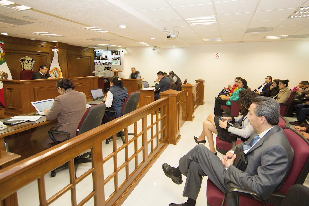 Vista panorámica en ángulo diagonal de una sala de audiencias iluminada, con un juez presidiendo al fondo y asistentes sentados en butacas guinda a la derecha separadas por una barandilla de madera.