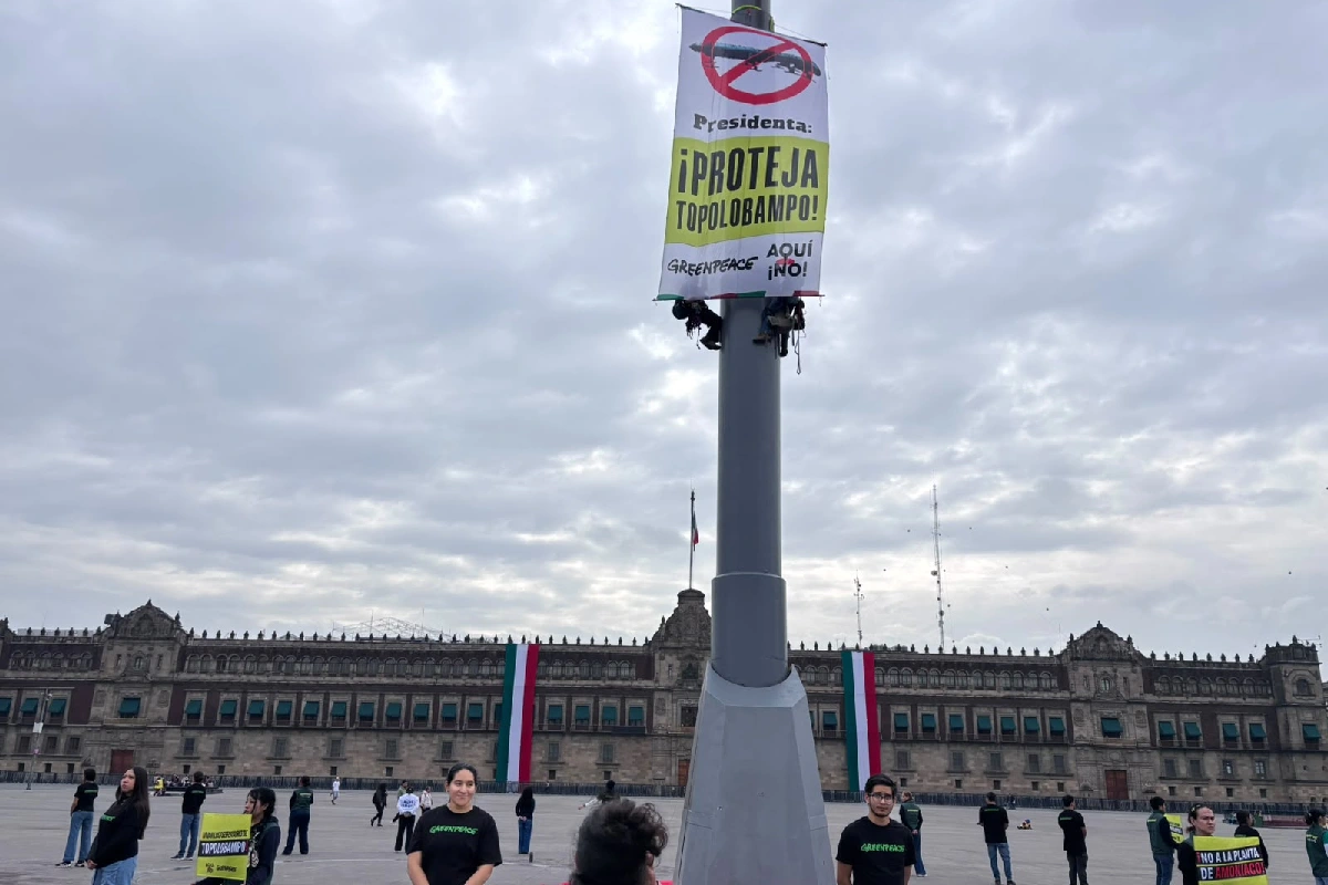 Plano medio de la base del asta bandera en el Zócalo de la Ciudad de México, donde activistas de Greenpeace cuelgan una lona con la leyenda "Presidenta: ¡PROTEJA TOPOLOBAMPO!", con manifestantes reunidos alrededor y Palacio Nacional al fondo.
