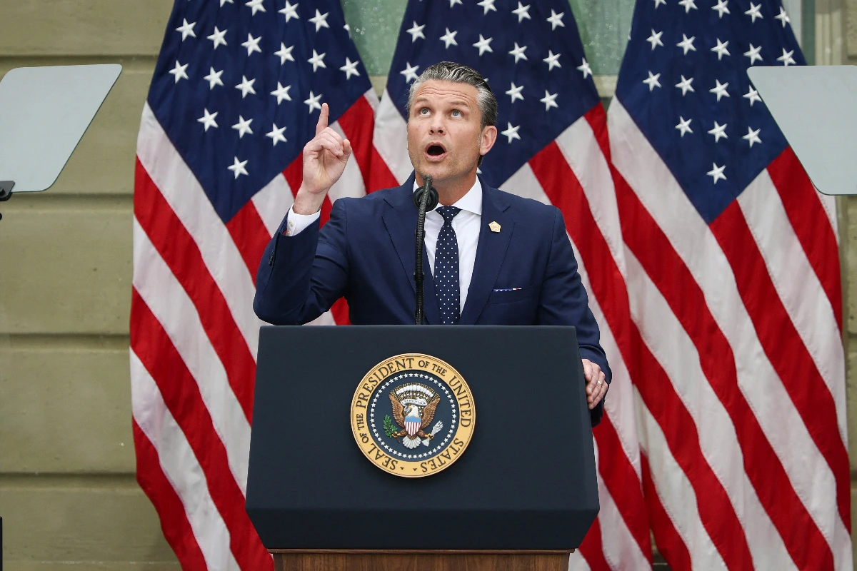 Pete Hegseth señalando con el dedo índice hacia arriba mientras habla detrás del pódium con el sello oficial del Presidente de los Estados Unidos, frente a tres banderas estadounidenses.