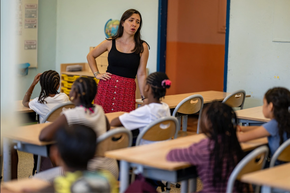 Profesora de pie vestida con blusa tirantes negra y falda roja impartiendo clase frente a un grupo de estudiantes sentados en sus bancas escolares.