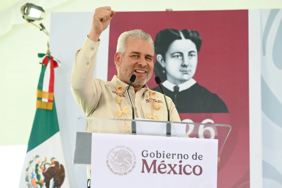 El gobernador Alfredo Ramírez Bedolla levantando el puño derecho con una sonrisa desde un pódium con la leyenda "Gobierno de México" y la bandera nacional al lado.