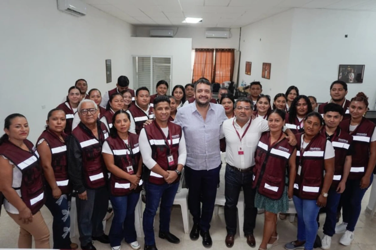 Andrés Manuel López Beltrán al centro sonriendo junto a un grupo numeroso de militantes y simpatizantes que visten chalecos guinda con reflejantes con el logo de Morena en las oficinas del partido en Tabasco.