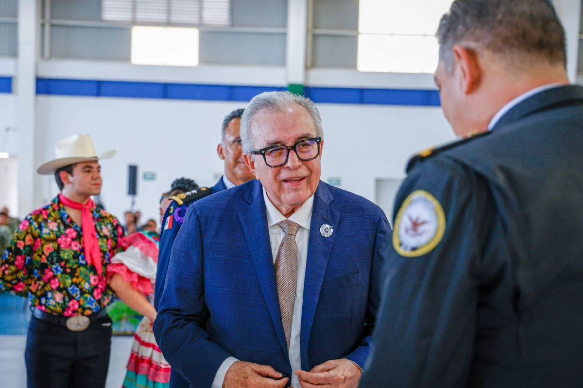 Fotografía de Rubén Rocha Moya, gobernador de Sinaloa, sonriendo y mirando a un oficial de policía de uniforme durante un evento público. Viste un traje azul y gafas. Al fondo, un joven con sombrero de vaquero y camisa floreada, y personas con trajes folclóricos.