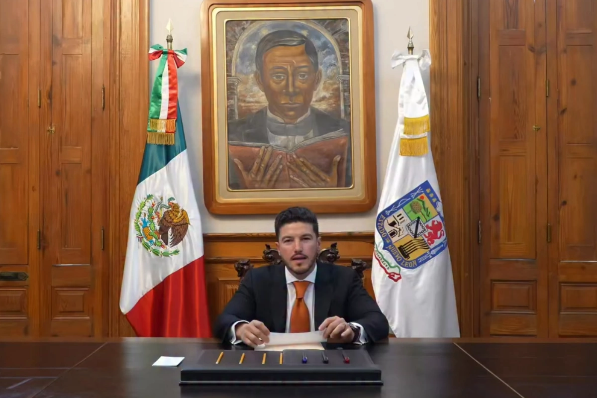El gobernador Samuel García sentado frente a su escritorio en su despacho institucional, flanqueado por la bandera de México y la de Nuevo León, mientras emite un mensaje oficial.