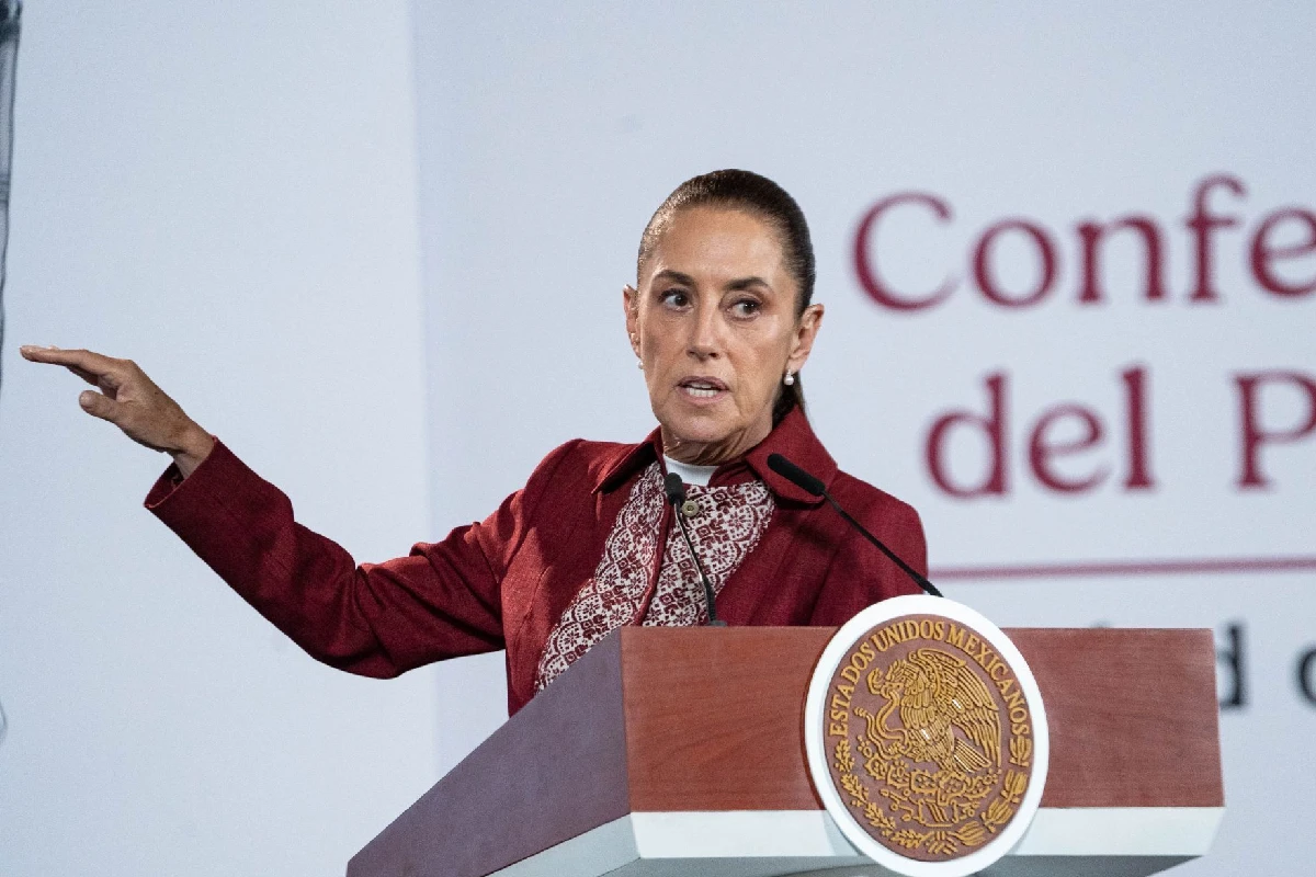 Claudia Sheinbaum de pie tras el pódium presidencial con el escudo de México, vistiendo un saco guinda y una blusa bordada artesanal mientras extiende el brazo derecho al hablar en la "Conferencia del Pueblo".