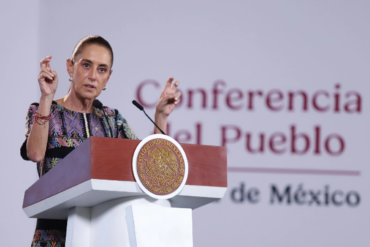 Claudia Sheinbaum de pie tras el pódium presidencial gesticulando con los dedos en forma de comillas, vistiendo un vestido oscuro con bordados artesanales multicolor ante la escenografía de la "Conferencia del Pueblo".