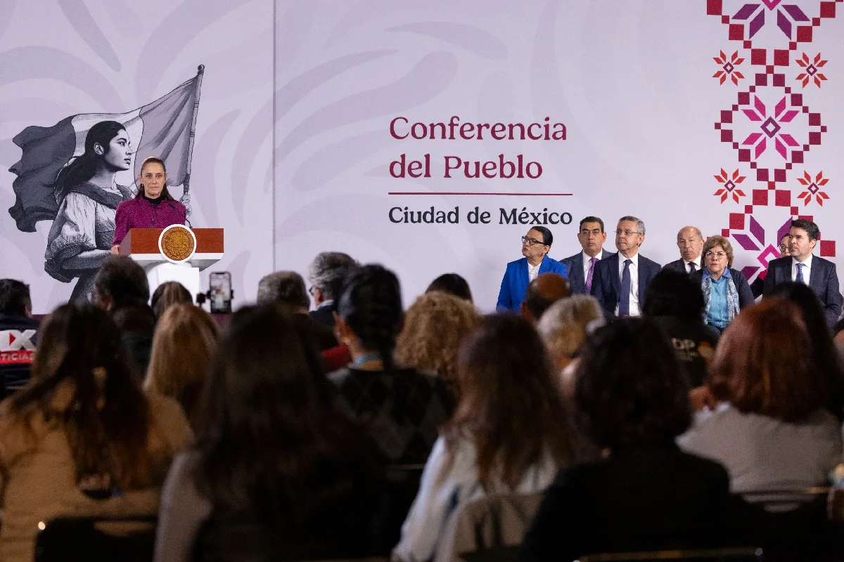 Claudia Sheinbaum encabezando la "Conferencia del Pueblo" desde el pódium, acompañada por miembros de su gabinete sentados a la derecha y frente a un grupo de periodistas en el salón.