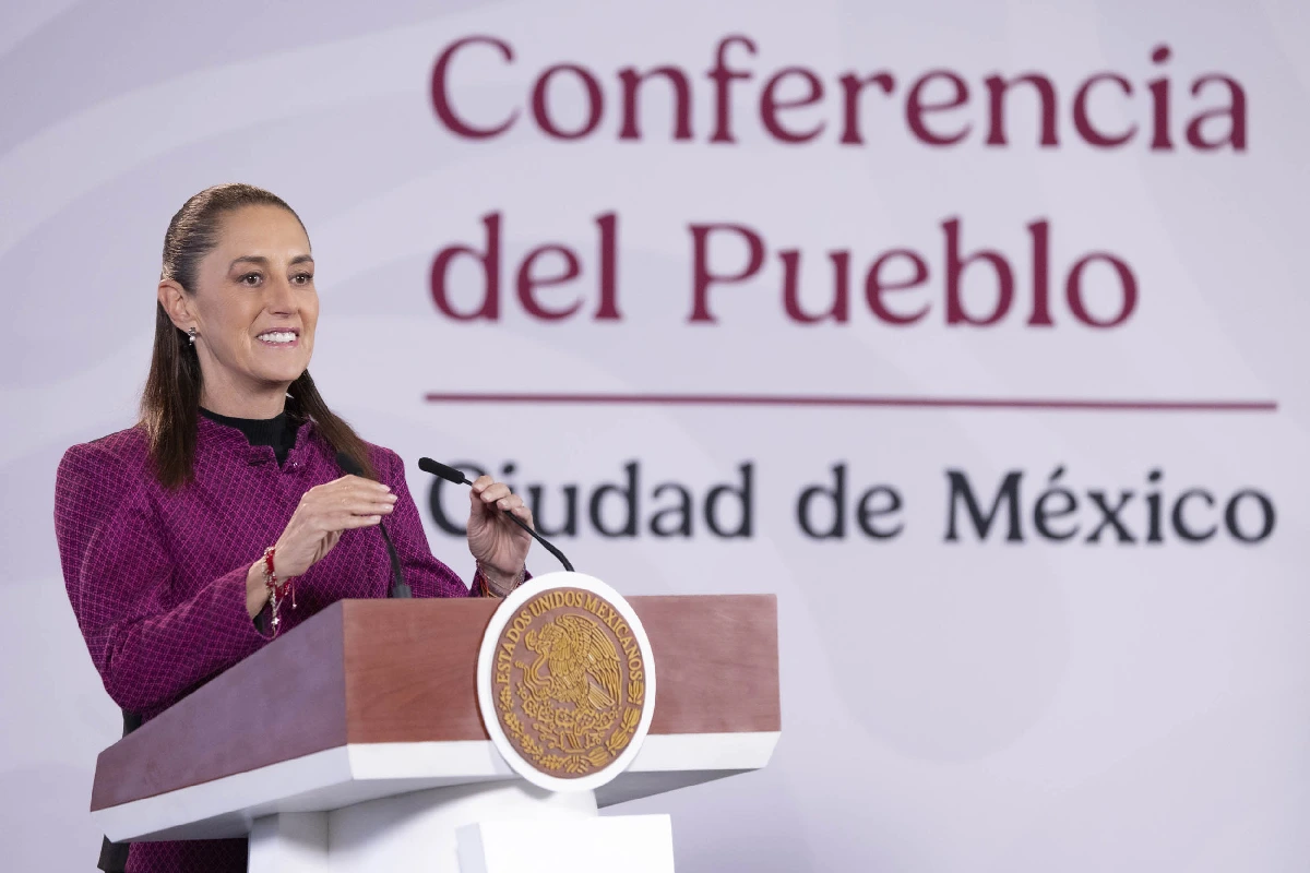 Claudia Sheinbaum sonriendo y gesticulando con las manos frente a los micrófonos en un pódium de madera con el sello institucional, con el fondo de la "Conferencia del Pueblo".