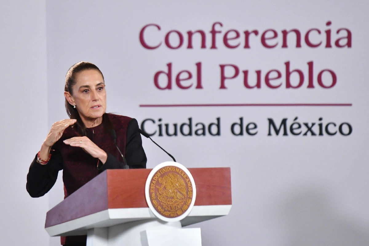 Claudia Sheinbaum de pie tras el pódium con el escudo de México, haciendo un gesto explicativo con ambas manos abiertas frente al letrero de la "Conferencia del Pueblo".