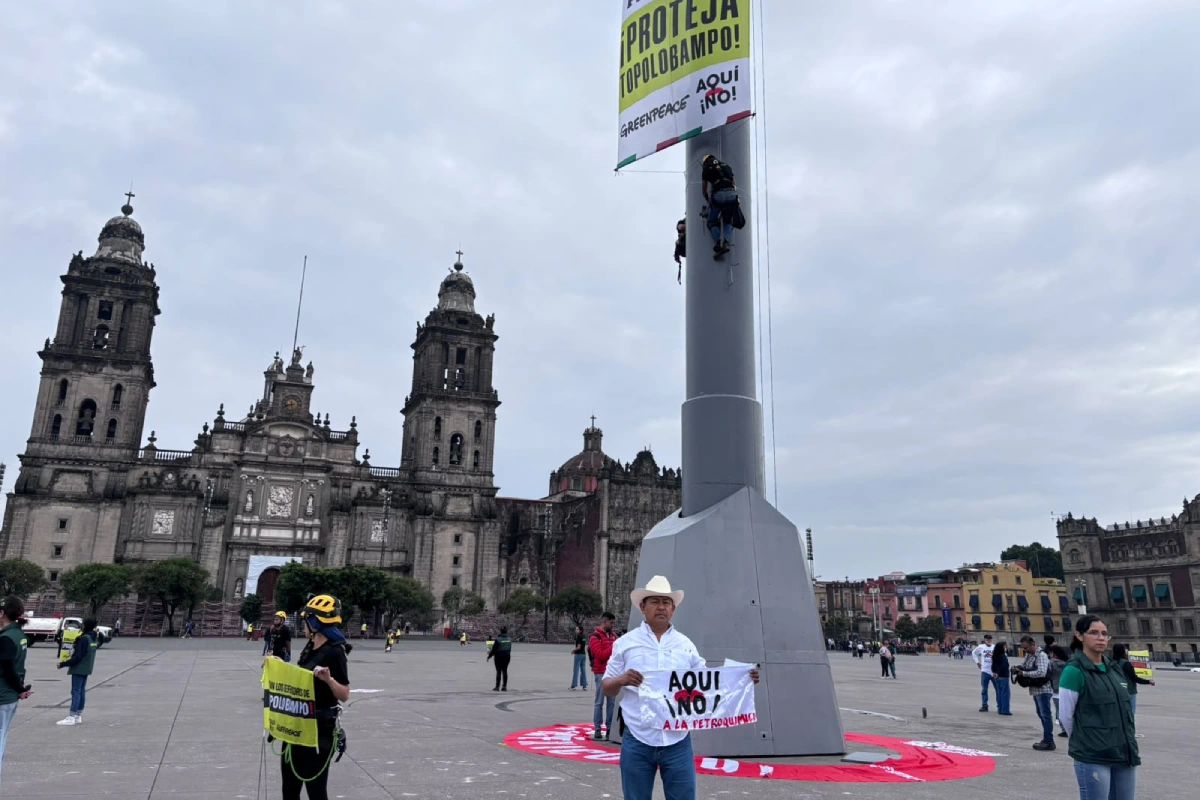 Manifestantes en el Zócalo de la Ciudad de México frente a la Catedral Metropolitana, con un hombre en primer plano sosteniendo el cartel "¡Aquí NO! A la petroquímica" y activistas de Greenpeace escalando el asta bandera para colgar una lona con la frase "¡Proteja Topolobampo!".