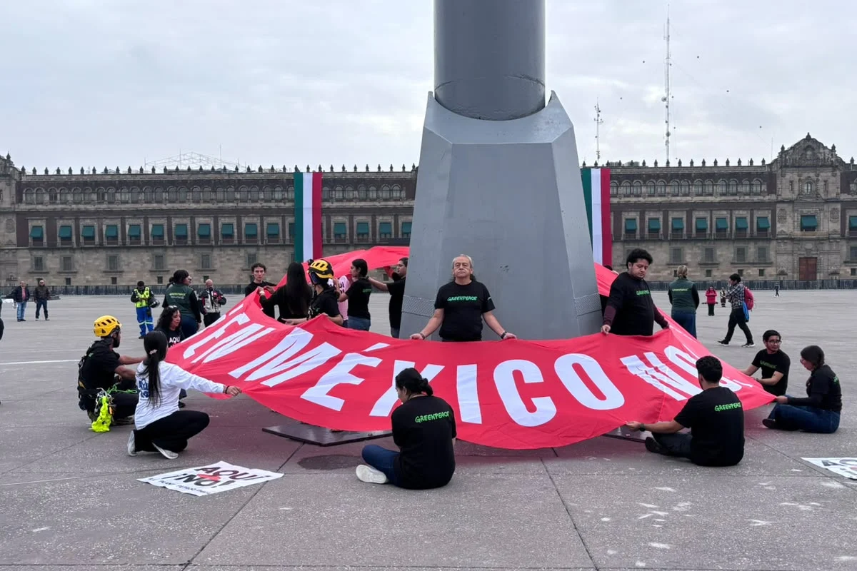 Activistas medioambientales de Greenpeace rodeando la base del asta bandera en el Zócalo de la Ciudad de México, desplegando una manta roja gigante con la leyenda "EN MÉXICO NO" frente a Palacio Nacional.