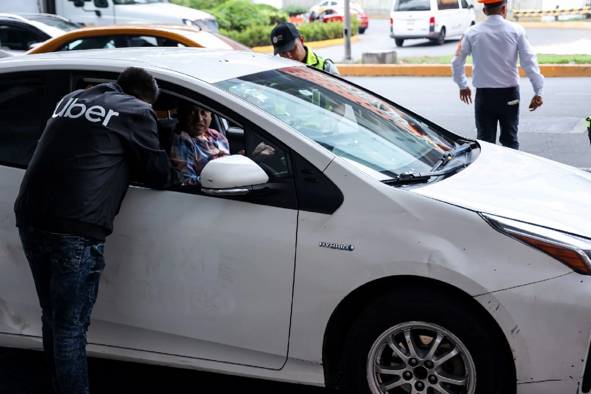 Un hombre vistiendo una chamarra con la palabra "Uber" inclinándose para hablar con el conductor de un automóvil híbrido blanco detenido en la vía pública, con inspectores de tránsito cerca.
