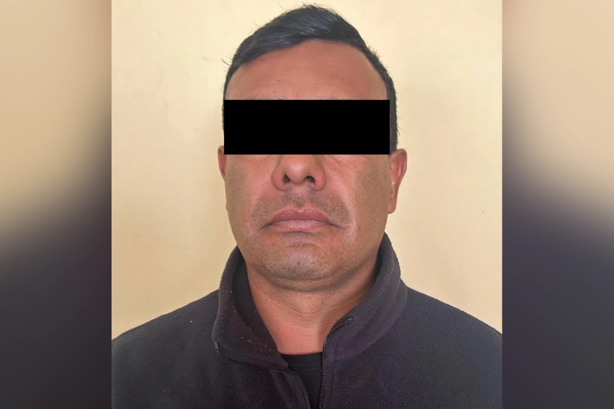 Fotografía de frente de un hombre adulto con cabello oscuro y corto, vistiendo una chamarra de lana negra con cuello alto, parado frente a una pared beige lisa, con los ojos censurados por una barra negra.